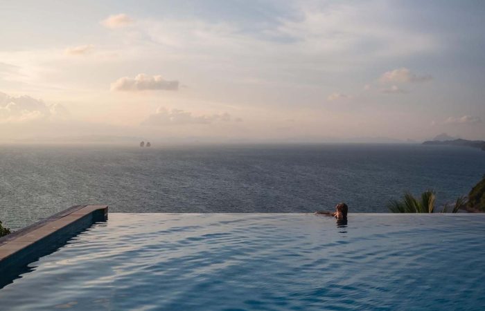 A young woman relaxing in infinity swimming pool and looking at a beautiful sunset and the sea view